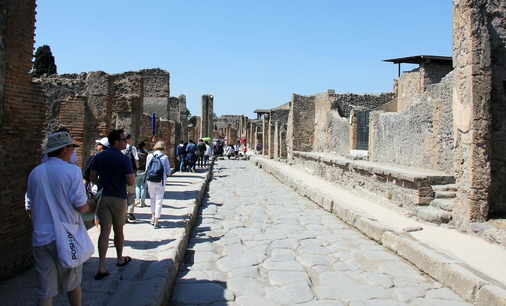 Back on the road - this time the Via Abbondanza. As you can see, Pompeii is very popular with sightseers. The tall stone pillar with the vertical groove
        up ahead is part of the water distribution system to fountains all over the town.