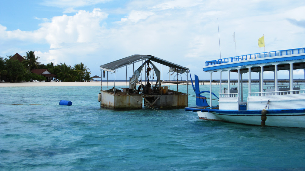 The rather ugly, rusting sand-pumping pontoon and its outflow pipe supported by a line of blue barrels. Room no 4 is behind the palm trees at the left of the photo.  (192k)