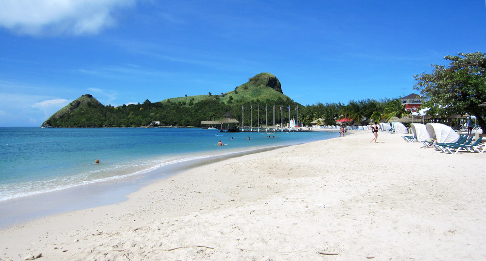 View from the far end of the Grande's beach, next to the breakwater, looking back towards Pigeon Island, with Signal Peak in the middle. Fort Rodney is on
        top of the lower, left-hand peak.  (126k)