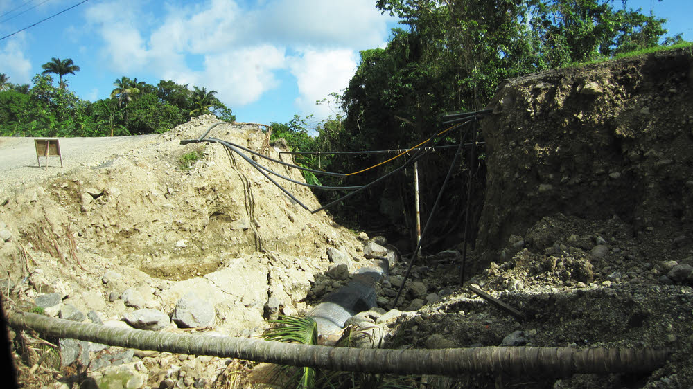 The same location. We've left the road, driven down the bank to the right of the road, and are bumping over the stream bed, looking back up to
        where the road was washed away. You can see the culvert at the bottom of the gulley, while water pipes normally buried at the side of the road are
        dangling over the hole. (173k)